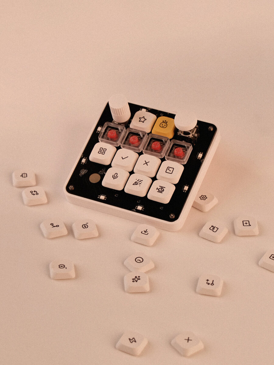 OpenMicroKbd with the stock keycap set installed — a star, a yellow crowned mascot, four clear caps, a grid, a check, a cross, a terminal prompt, a microphone, a party popper and a robot — surrounded by loose spare keycaps