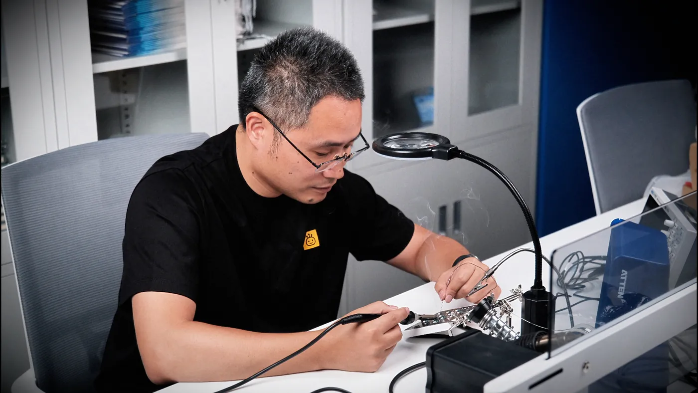 Hand-soldering the board at an electronics workbench, with magnifier, iron and board vise in frame