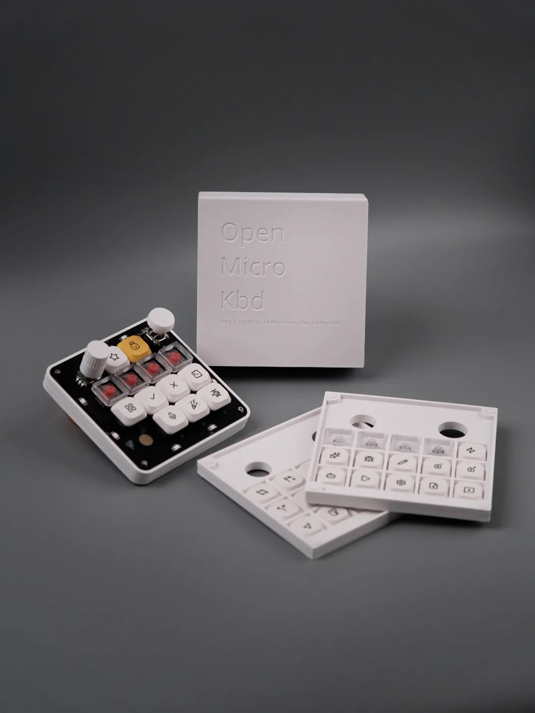 The assembled board in its white printed case, beside the embossed Open Micro Kbd packaging box and two printed trays of alternate keycaps