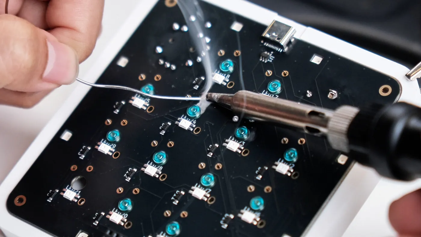Macro view of the board's underside during hand assembly: a soldering iron and solder wire at one joint, surrounded by the teal reverse-mount LED packages silkscreened LED17 through LED29